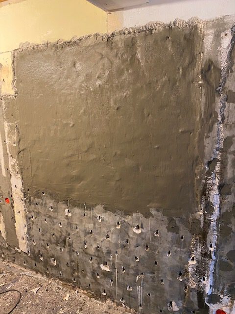 Eine teilweise verputzte Wand mit rauen, unbearbeiteten Kanten und sichtbaren Bohrlöchern im unteren Bereich, die auf laufende Bau- oder Renovierungsarbeiten im Injektionsverfahren zur effektiven Bauwerksabdichtung hinweisen.