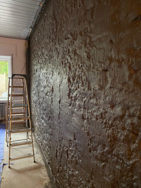 Eine metallene Trittleiter steht neben einer rauen, frisch verputzten Wand - Teil der laufenden Bautenschutzarbeiten - in einem Raum mit weißer Decke und natürlichem Licht, das aus einem Fenster auf der linken Seite kommt.
