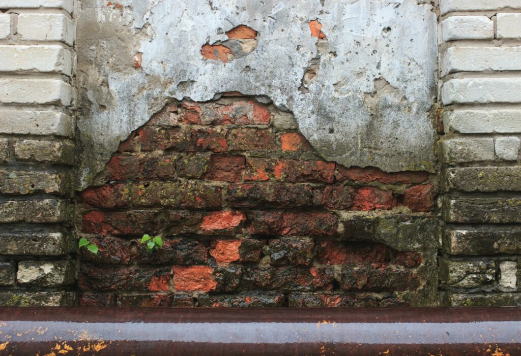 Freigelegte rote Ziegel und abgenutzter Mörtel zeigen sich durch einen bröckelnden Putzfleck an einer alten Mauer, die von weißen Ziegeln umrahmt ist - ein Beispiel dafür, wie Bautenschutz und Abdichtung von Mauerwerk einen weiteren Verfall verhindern könnten.